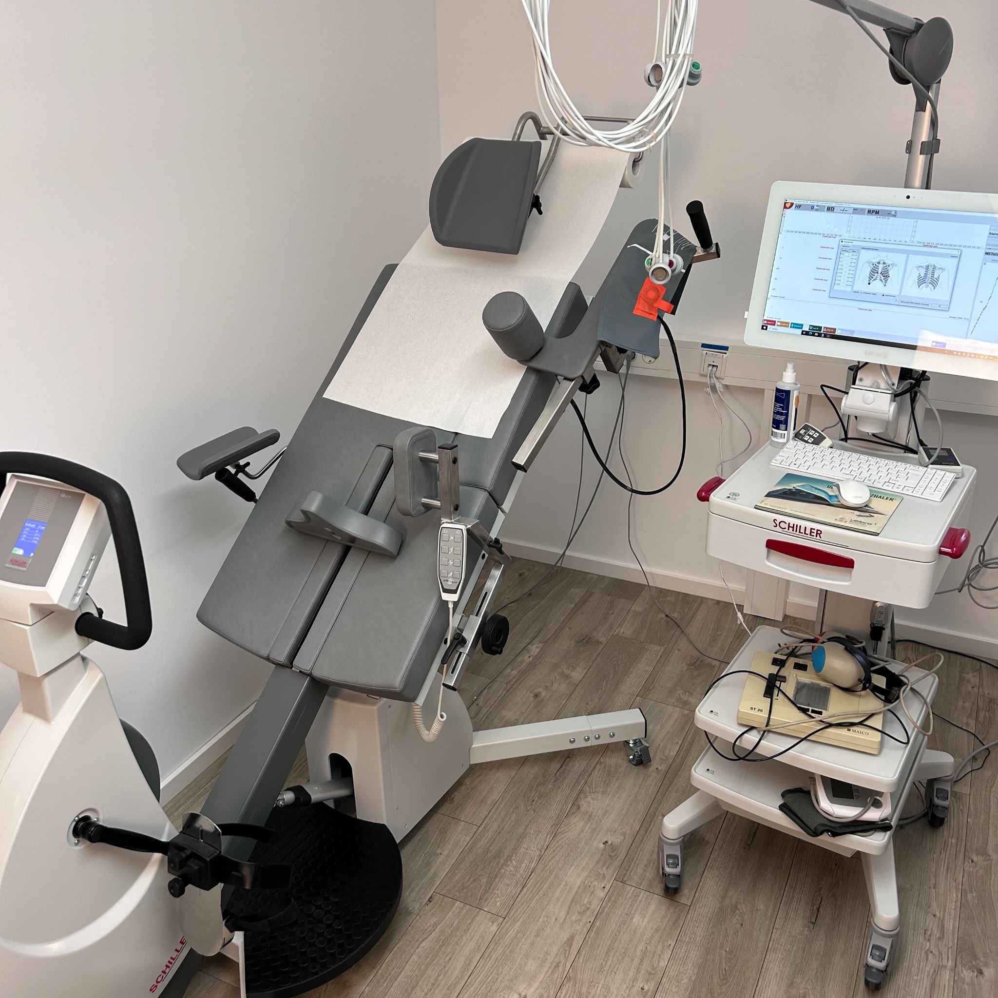 Ein älterer Mann sitzt auf einem medizinischen Stuhl und unterzieht sich einer Belastungs-EKG-Untersuchung. Er ist mit Elektroden verbunden, die auf seiner Brust angebracht sind, während eine Ärztin seinen Blutdruck misst und ein weiterer Arzt die Untersuchung überwacht.